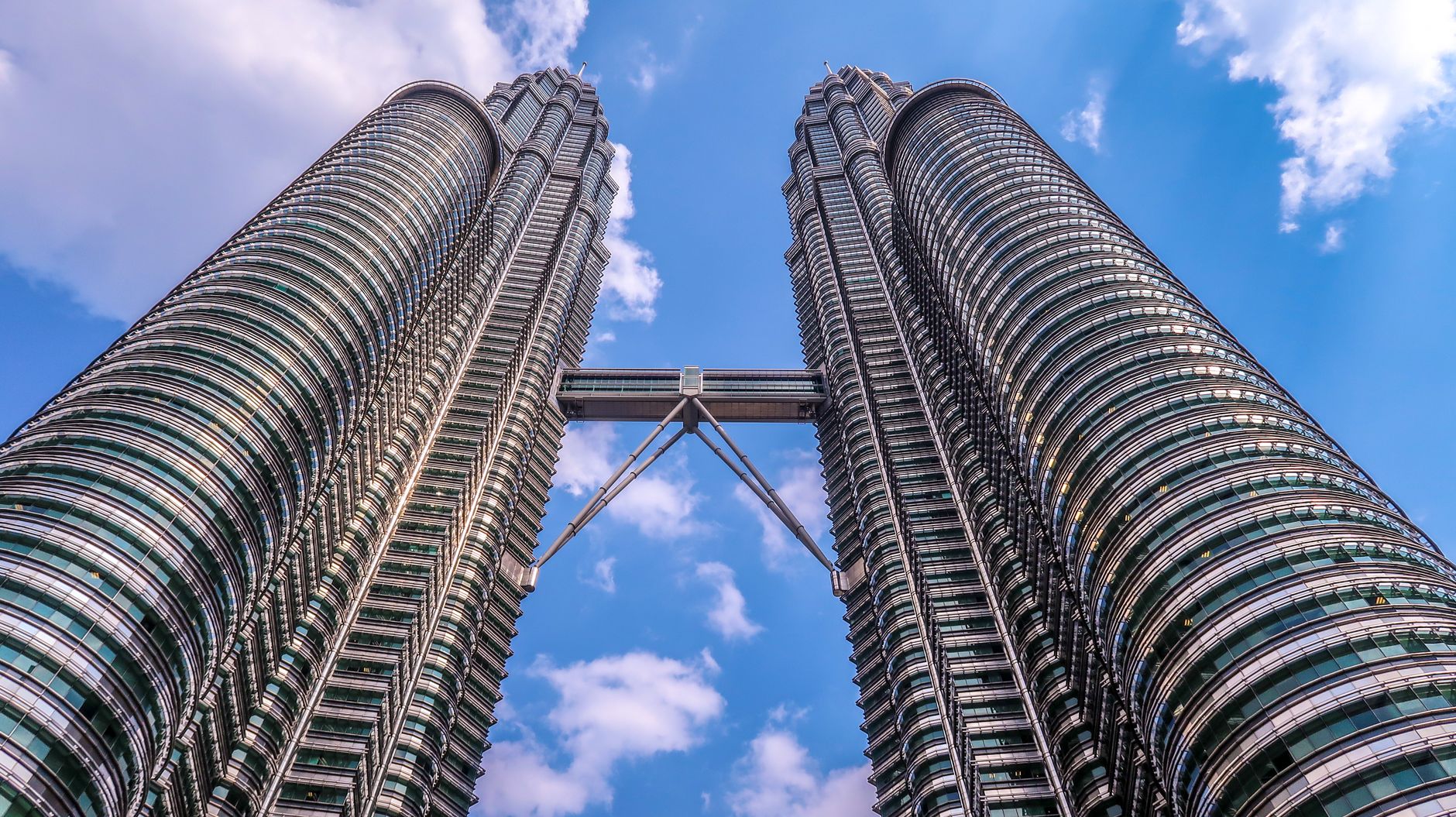 Low-angle view of the Petronas Twin Towers in Kuala Lumpur, Malaysia, joined by the skybridge under a blue sky with scattered cloud.