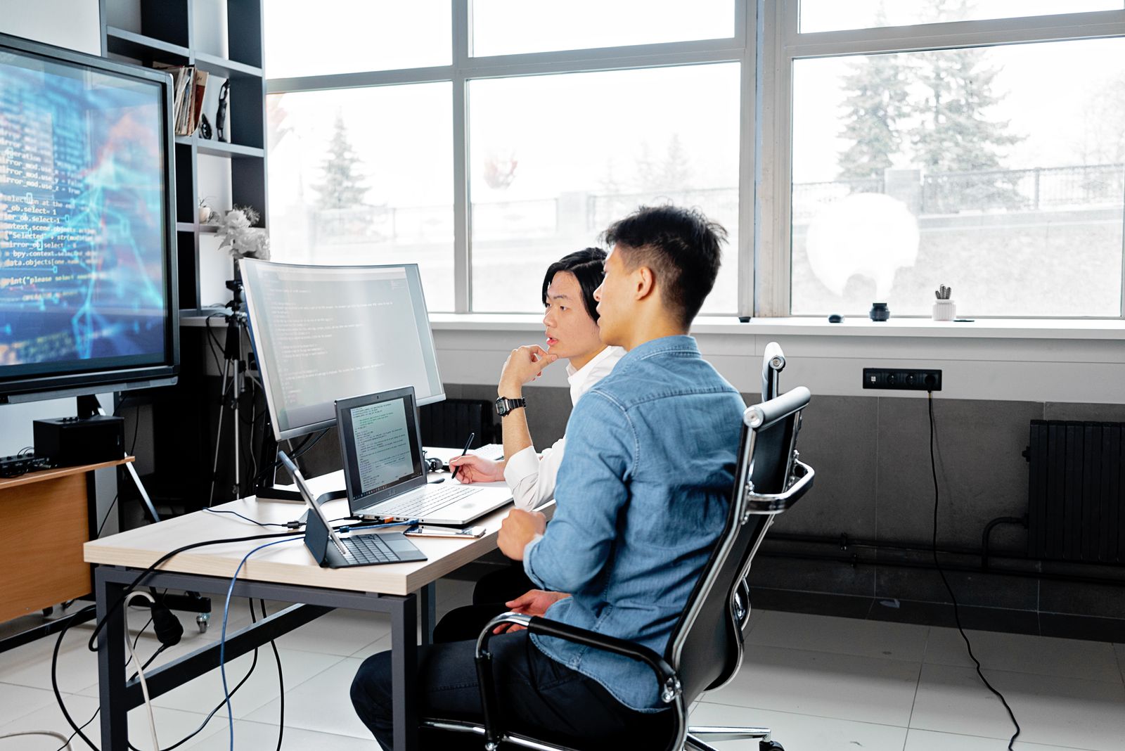 Two colleagues sitting at a shared desk in a bright office, reviewing code on a laptop and monitors.