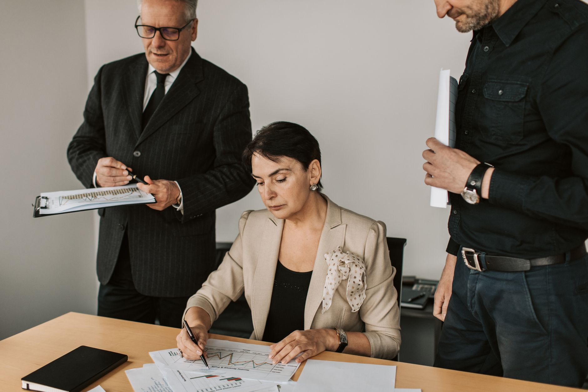 A senior professional reviews and signs a document at a desk while two colleagues stand beside her holding papers and a clipboard.