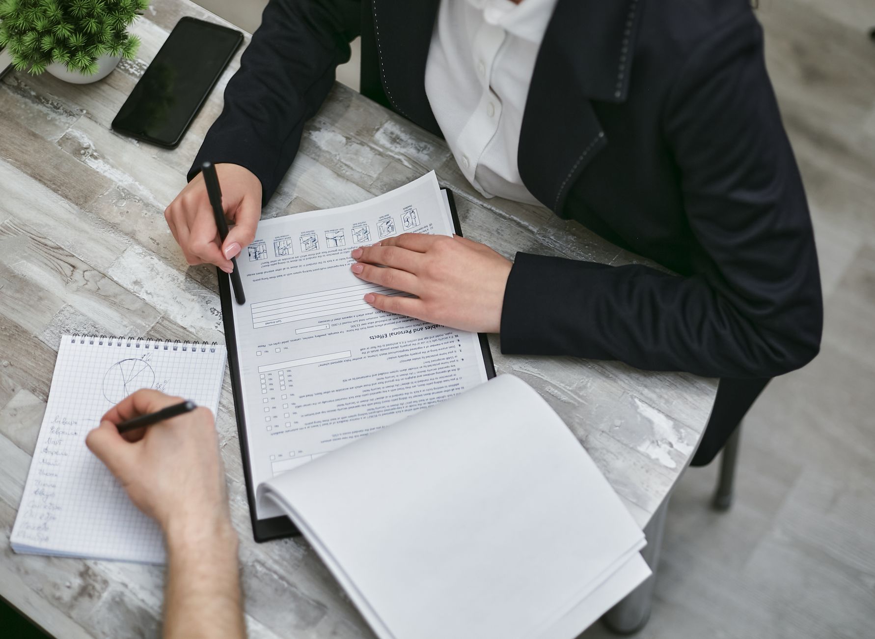 Two colleagues sit at a desk reviewing printed documents and taking notes during a meeting.