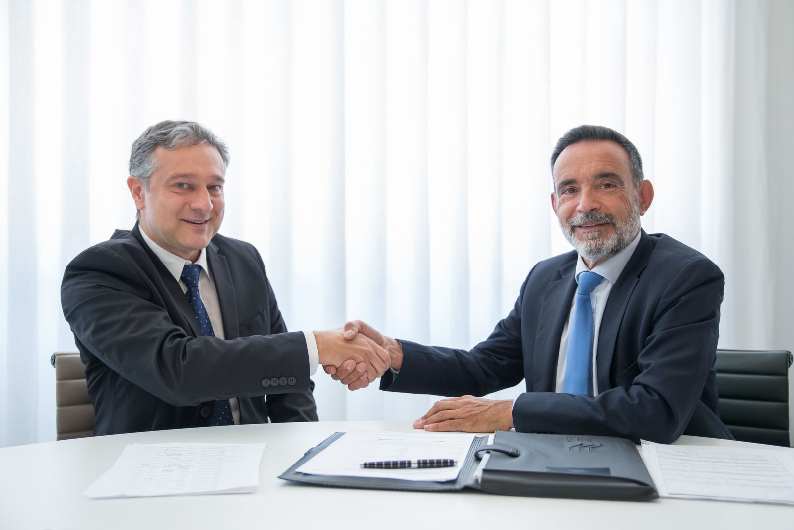 Two business people shaking hands across an office table with documents, representing a supplier relationship and its contractual terms